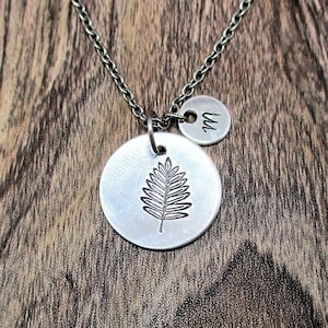 May include: A silver necklace with a leaf charm and a small disc charm with the letter "W" engraved on it. The necklace is on a wooden surface.