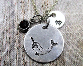 May include: A silver-toned necklace featuring a circular pendant stamped with a black narwhal illustration, a smaller circular charm stamped with the letter M, and a small round dark red gemstone charm suspended from a delicate metal chain.