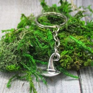 May include: A silver-colored keychain featuring a witch hat charm. The charm is attached to a chain and a keyring. The keychain is set against a backdrop of vibrant green moss.