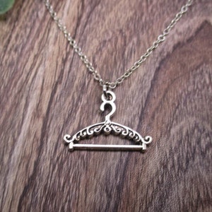May include: Silver-toned necklace featuring a detailed miniature clothes hanger pendant. The pendant has an ornate, decorative design. The necklace hangs on a silver chain, set against a rustic wooden background with a touch of greenery.