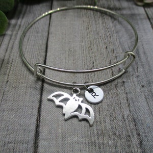 May include: A silver-toned bangle bracelet with a bat charm and a small round charm with the letter "R". The bat charm is cut out and has a detailed wing design. The bracelet is displayed on a gray wooden surface.