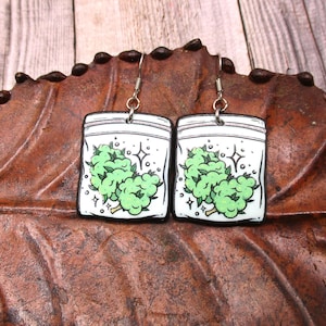 May include: A pair of silver dangle earrings with a white background and a green marijuana leaf design. The earrings are shaped like a small plastic bag with a green marijuana leaf inside.