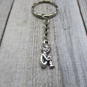 May include: Silver-toned keychain featuring a small, detailed figurine of a seated goblin or imp. The figure is attached to a chain and a keyring. The background is a light gray wooden surface.