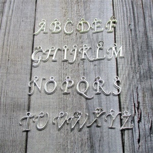 May include: Silver alphabet letter charms, A through Z, on a wooden background. Each letter is a separate charm.