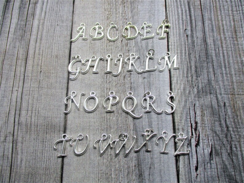 May include: Silver alphabet letter charms, A through Z, on a wooden background. Each letter is a separate charm.