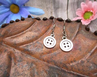 May include: Silver button earrings with four holes on each button. The earrings are hanging from a brown leaf.