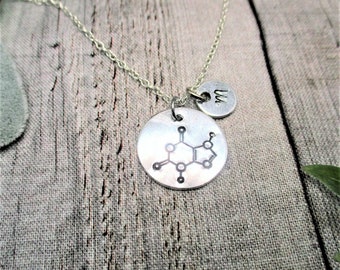 May include: A silver-toned necklace featuring a circular pendant engraved with a black chemical structure diagram and a smaller circular charm stamped with the letter M, suspended from a delicate silver-toned metal chain link necklace.