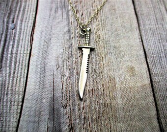 May include: A silver-toned metal pendant shaped like a survival knife with a serrated edge and a textured handle, suspended from a matching thin metal chain. The pendant features a pointed blade and a crossguard, hanging vertically from a small jump ring.
