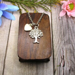 Puede incluir: Un collar de plata con un colgante de &aacute;rbol de la vida y un peque&ntilde;o dije en forma de coraz&oacute;n. El collar se muestra en un soporte de madera.
