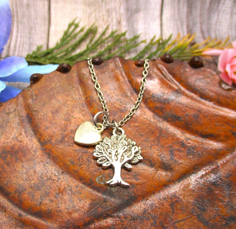 Puede incluir: Un collar de plata con un dije de &aacute;rbol y un peque&ntilde;o dije de coraz&oacute;n. El collar est&aacute; sobre una superficie marr&oacute;n texturizada.