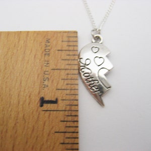 May include: Silver necklace with a broken heart pendant that says "Mother" on one half and has two small hearts on the other half. The necklace is measured against a ruler.