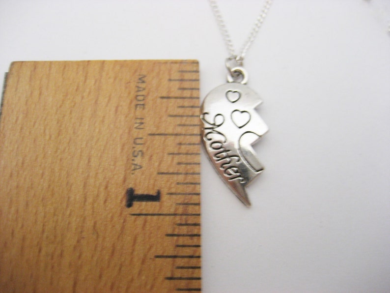 May include: Silver necklace with a broken heart pendant that says "Mother" on one half and has two small hearts on the other half. The necklace is measured against a ruler.