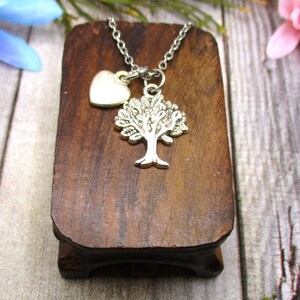 Puede incluir: Un collar de plata con un colgante de &aacute;rbol y un peque&ntilde;o dije en forma de coraz&oacute;n. El collar se muestra en un soporte de madera.