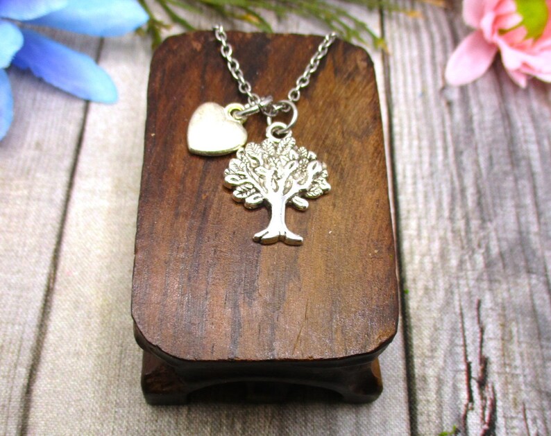 Puede incluir: Un collar de plata con un colgante de &aacute;rbol y un peque&ntilde;o dije en forma de coraz&oacute;n. El collar se muestra en un soporte de madera.