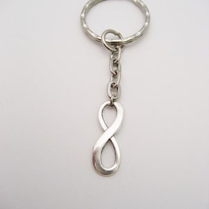 May include: Silver-toned keychain featuring an infinity symbol charm. The keychain includes a split ring, a chain, and a stylized figure eight charm. The metal has a slightly aged appearance, and the background is plain white.