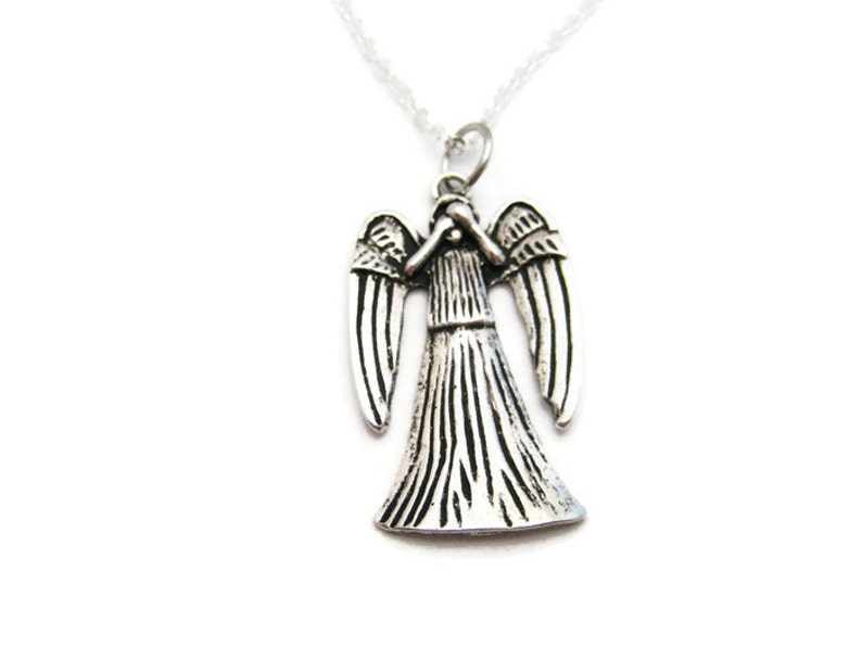 May include: A silver angel pendant necklace with a detailed design. The angel is depicted with wings outstretched and its head bowed. The necklace is on a silver chain.