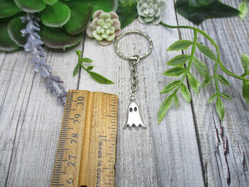 May include: A silver ghost keychain with a silver key ring. The ghost has two black eyes and a white body.