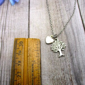 Puede incluir: Un collar de plata con un dije de &aacute;rbol y coraz&oacute;n. El collar est&aacute; sobre una superficie de madera con una regla para referencia de tama&ntilde;o.
