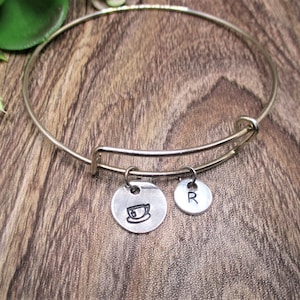 May include: A silver-colored bangle bracelet with two small, round charms. One charm features a teacup design, and the other has the letter "R". The bracelet is displayed on a wooden surface.