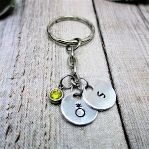 May include: A silver keychain with a round key ring, chain, and three charms. The charms include a small, round, silver charm with the letter "S", a silver charm with a symbol, and a small, round, yellow-green gemstone.
