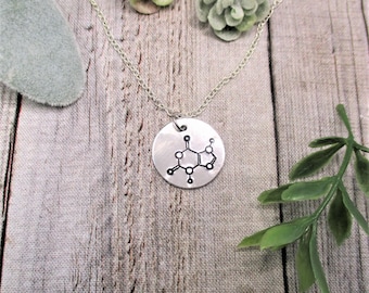May include: A silver-toned circular pendant necklace featuring a black engraved chemical structure of a caffeine molecule. The pendant hangs from a delicate silver-toned chain, showcasing a polished metallic finish with clear, dark etched lines.