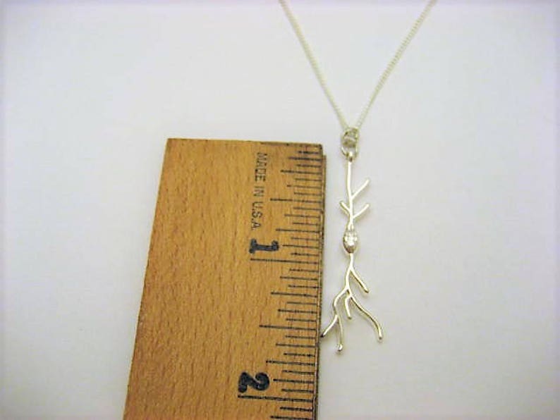 Puede incluir: Un collar de plata con un delicado colgante de rama. El colgante presenta una peque&ntilde;a piedra transparente en el centro de la rama. El collar mide aproximadamente 2,5 cm de largo.