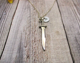 May include: A silver-toned metal necklace featuring a pendant shaped like a serrated knife and a small circular charm stamped with the letter M. The pendant and charm hang from a thin, matching silver-toned metal chain link necklace.