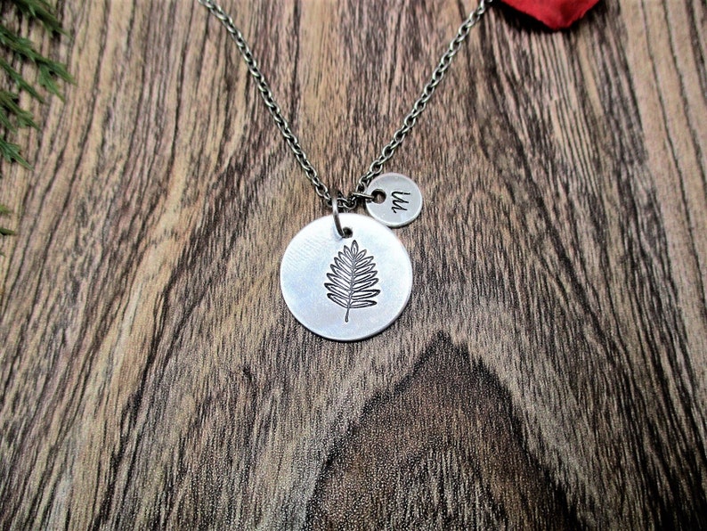 May include: A silver necklace with a leaf charm and a small disc charm with the letter "W" engraved on it. The necklace is on a wooden surface.