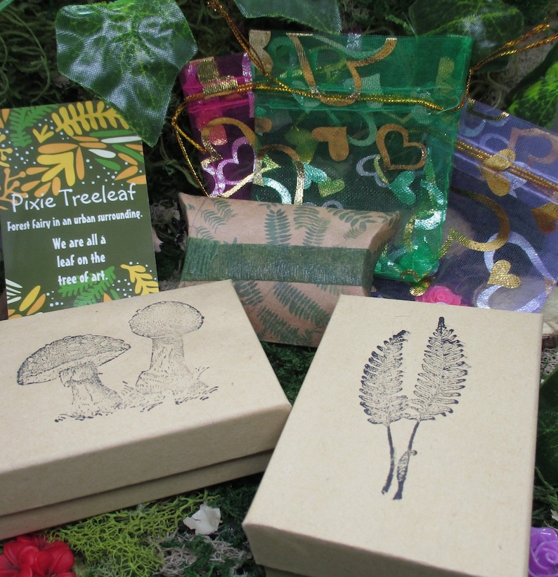 May include: Two small brown boxes with different designs. One box has a mushroom illustration and the other has a fern illustration. The boxes are surrounded by green moss and a card with the text "Pixie Treeleaf Forest Fairy in an urban surrounding. We are all a leaf on the tree of art."