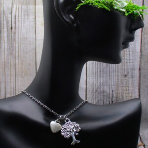 Puede incluir: Un collar de plata con un dije de &aacute;rbol y un dije de coraz&oacute;n. El collar est&aacute; en un maniqu&iacute; negro con una corona floral verde.