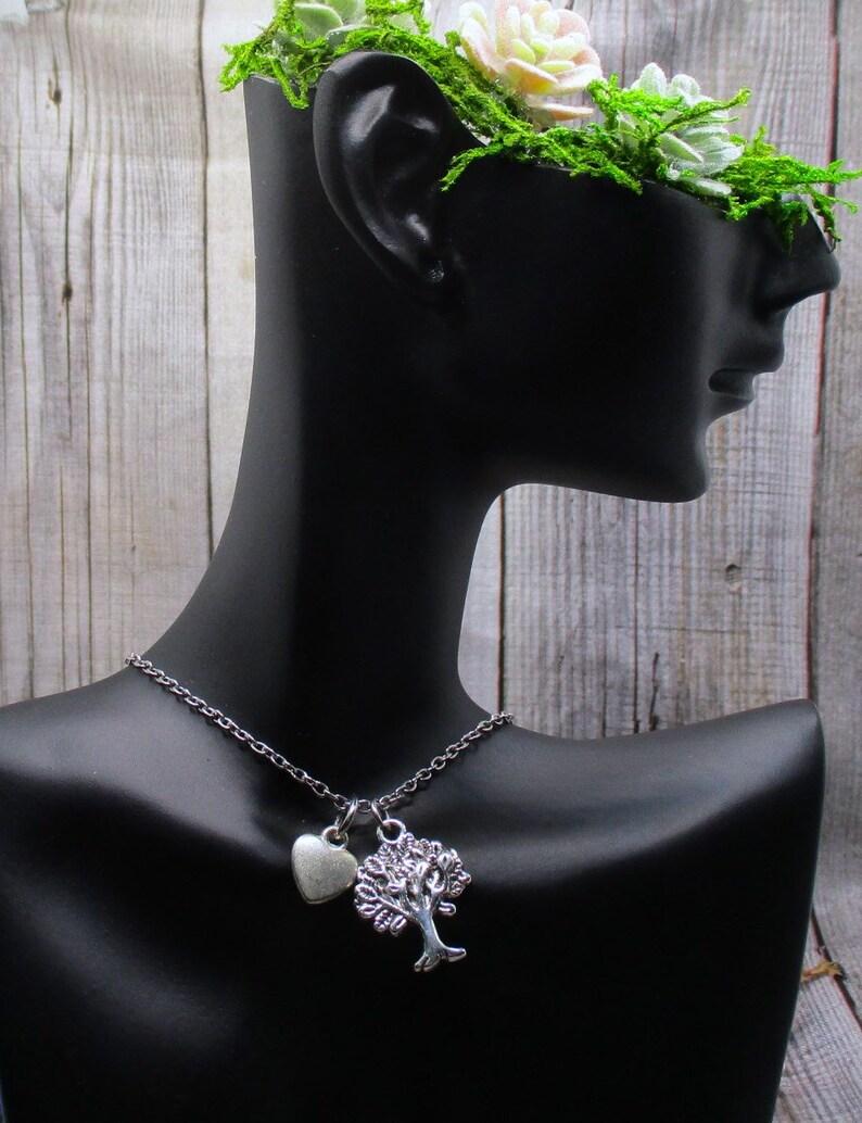 Puede incluir: Un collar de plata con un dije de &aacute;rbol y un dije de coraz&oacute;n. El collar est&aacute; en un maniqu&iacute; negro con una corona floral verde.