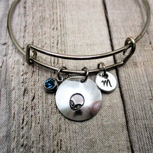 May include: A silver expandable bangle bracelet with a charm featuring an alien face, a blue crystal charm, and a charm with the letter "M" engraved on it.
