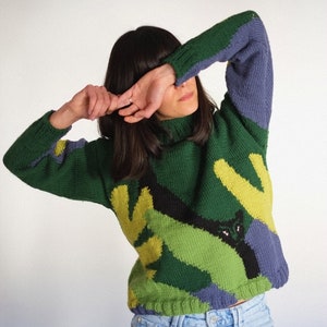 Op de afbeelding: Een groene, gele en paarse gebreide trui met een zwart kattenontwerp. De trui heeft een hoge hals en lange mouwen.