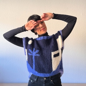 Könnte beinhalten: Ein blauer und weißer, gestrickter, ärmelloser Pullover mit geometrischem Muster. Der Pullover hat einen weißen Kreis, einen Baum und ein weißes Rechteck mit einem schwarzen Rechteck darin.