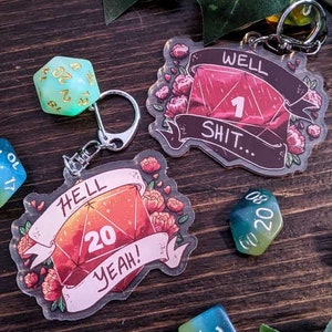 May include: Two acrylic keychains featuring dice with floral designs. One keychain has a red die with the number 20 and the text "HELL YEAH!" The other keychain has a red die with the number 1 and the text "WELL SHIT..."