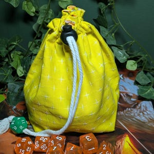 Könnte beinhalten: Ein leuchtend gelber Stoffbeutel mit Kordelzug und weißem Kreuzstichmuster, verschlossen mit einem schwarzen Kunststoff-Kordelstopper und weißem Seil. Davor liegen mehrere durchscheinende orangefarbene Polyederwürfel und ein grüner zwanzigseitiger Würfel.