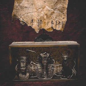 May include: A wooden box with a brown, straw-like material inside. Three small, silver-colored figurines are inside the box. The figurines are of an Egyptian pharaoh, a woman with a headdress, and a jackal. A piece of parchment with hieroglyphics is hanging above the box.