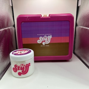 Puede incluir: Un tarro blanco con un logotipo rosa y rojo "The Stuff" se encuentra frente a un recipiente rosa estilo fiambrera. El recipiente tiene un diseño de bloques de color con las palabras "No Artificial Ingredients".