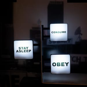 May include: Three illuminated square signs with the words "STAY ASLEEP", "CONSUME", and "OBEY" in a dark setting. The signs are white with black text. The background features a blurred cityscape.