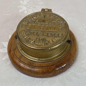 Puede incluir: Un medidor de agua antiguo con una parte superior de latón y una base de madera. La parte superior de latón está grabada con las palabras "Neptune Meters Co." y "Water Meter New York". El número de serie "5728911" también es visible. La base de madera es circular y tiene un tono marrón cálido.
