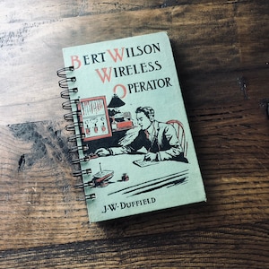 May include: A green spiral bound notebook with the title "Bert Wilson Wireless Operator" and the author "J-W. Duffield". The cover features an illustration of a man sitting at a desk with a radio and other equipment.