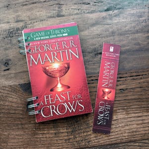 May include: A spiral-bound book and bookmark featuring the cover art for "A Feast for Crows" by George R.R. Martin. The book cover is red with a chalice illustration and the text "Game of Thrones" and "New York Times Bestseller".