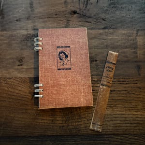 May include: A spiral-bound notebook with a textured, reddish-brown cover featuring a vintage illustration of a woman in a nurse's cap. Beside it lies a narrow, aged book titled "Cherry Ames at Spencer."