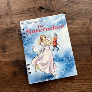 Peut inclure: Un carnet à spirales avec la couverture de "Casse-Noisette" d'un Petit Livre d'Or. L'illustration montre une femme en robe rose et un soldat en uniforme rouge sur un fond bleu étoilé.