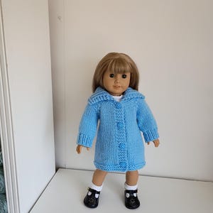 May include: A doll wearing a light blue knitted coat with a hood and button closures. The doll is also wearing black shoes with white socks. The coat is a solid color and appears to be handmade.