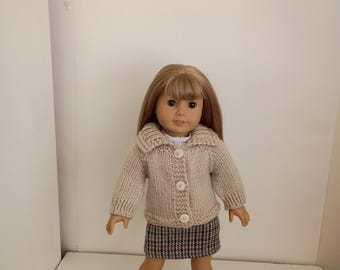 May include: A doll with shoulder-length blonde hair and bangs, wearing a beige knit cardigan with three white buttons, a white shirt underneath, a grey, black, and red plaid skirt, white socks, and black Mary Jane shoes with floral cutouts.