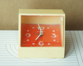 May include: A vintage cream-colored alarm clock with a red face and black hands. The clock has a clear plastic cover and the numbers 3, 6, 9, and 12 are printed on the face.