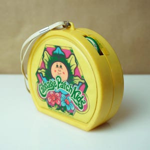 May include: A vintage yellow Cabbage Patch Kids radio with a white strap. The radio features a colourful graphic of a Cabbage Patch Kid and the brand name. The tuning dial is green.