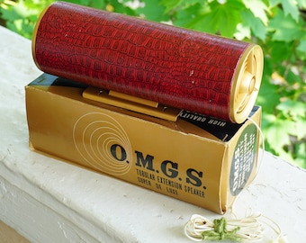 May include: A red tubular speaker with a gold trim and a crocodile-embossed finish. The speaker is sitting on a gold box with the text "O.M.G.S. TUBULAR EXTENSION SPEAKER SUPER DE LUXE" printed on it.