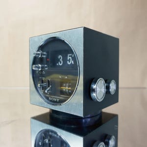 May include: A vintage black and silver Sony flip clock radio. The clock displays the time as 3:50. The clock has a square shape with a circular face and two silver knobs on the side. The word "SONY" is printed on the face.